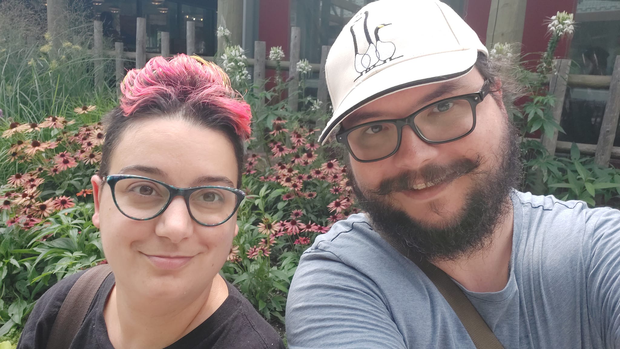 A man short hair and bushy facial hair sits next to a woman with a short rainbow mohawk in front of pink flowers